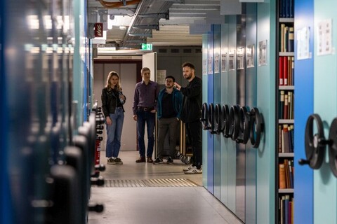 Vier Personen werden durch die Universitätsbibliothek im Westflügel geführt. Links und rechts blaue Regale mit Büchern.