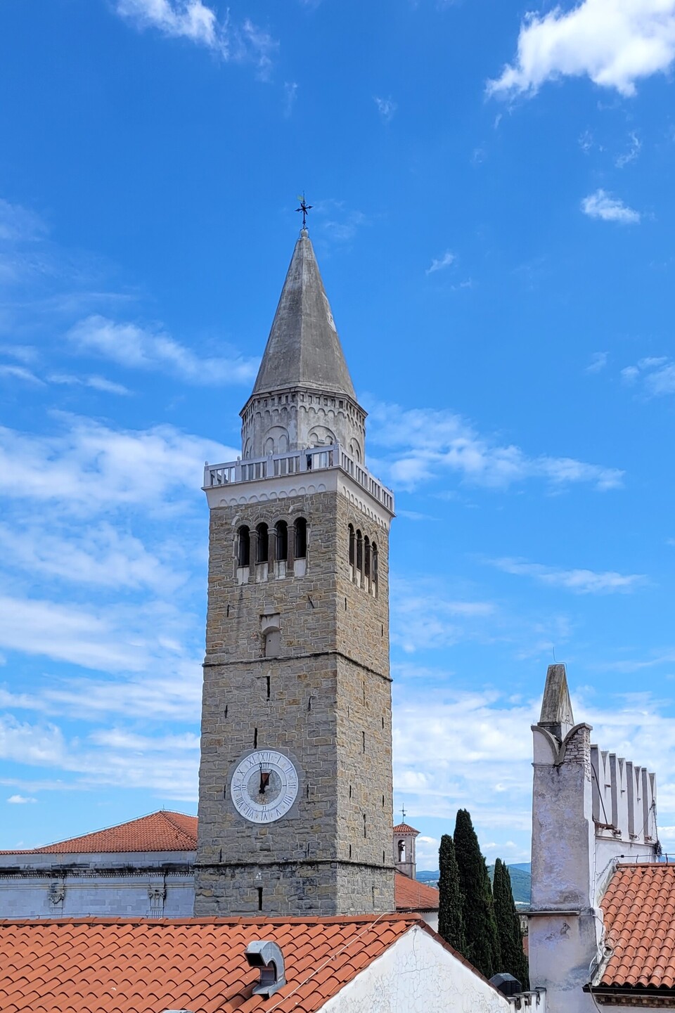 Ein steinerner Uhrturm mit spitzem Dach ragt vor einem strahlend blauen Himmel empor. Im Vordergrund sind rote Ziegeldächer und ein weißes Gebäude zu sehen.