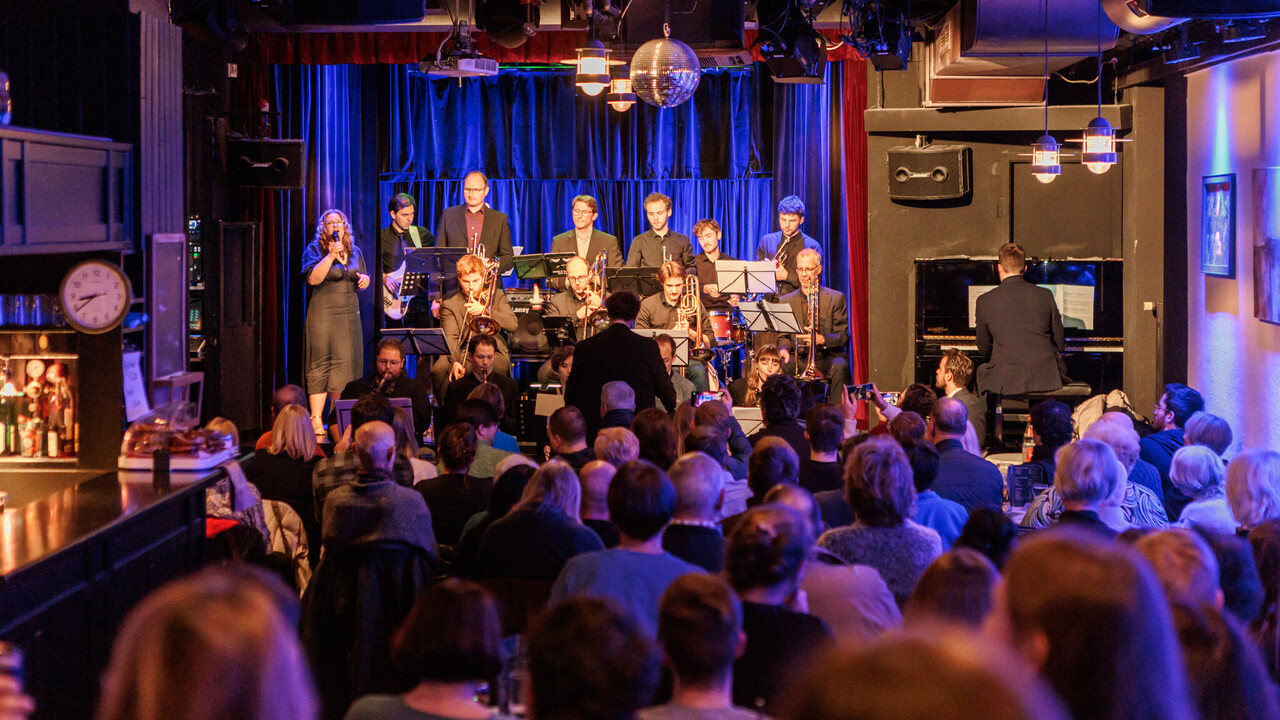 Eine Bigband spielt auf einer Bühne. Im Vordergrund sitzt Publikum, das von hinten zu sehen ist.