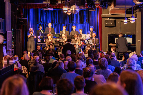 Eine Bigband spielt auf einer Bühne. Im Vordergrund sitzt Publikum, das von hinten zu sehen ist.