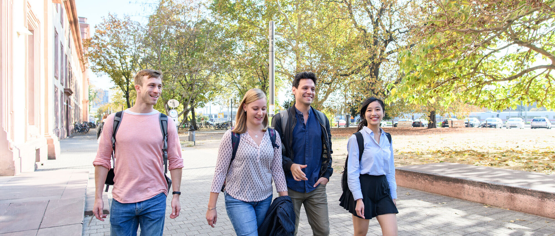 Vier Studierende laufen lachend auf einem Weg vor dem Schloss