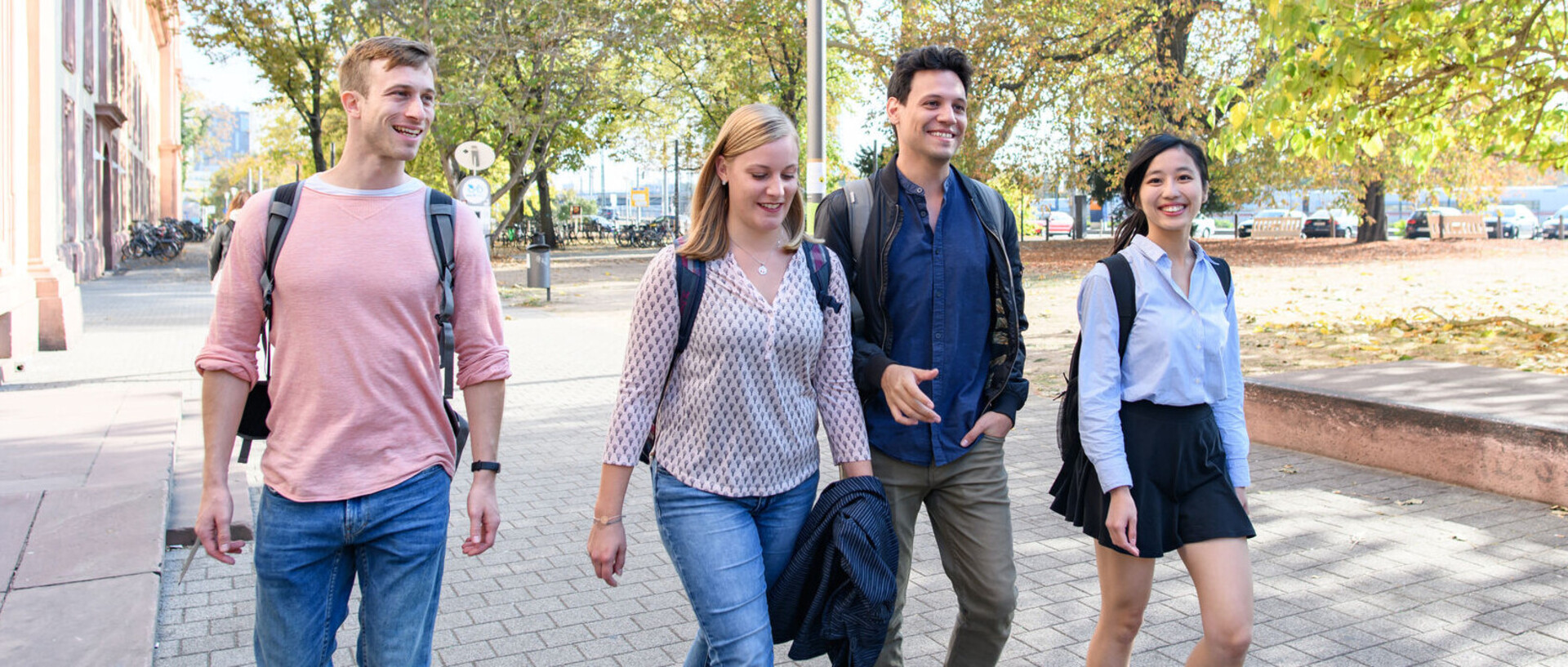 Vier Studierende laufen lachend auf einem Weg vor dem Schloss
