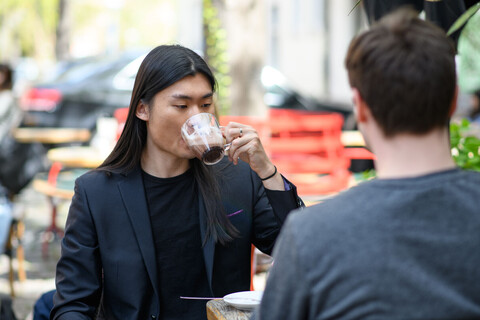 Ein Student mit glatten langen schwarzen Haaren und Jacket trinkt Kaffee aus einem Glas. Er sitzt draußen in einem Café.