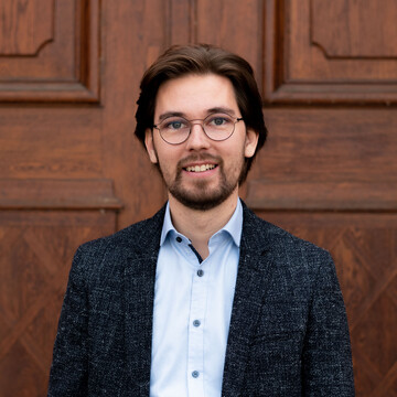 Julian Klix. Er trägt ein weißes Hemd und eine schwarze Jacke und steht vor einer Holztür.