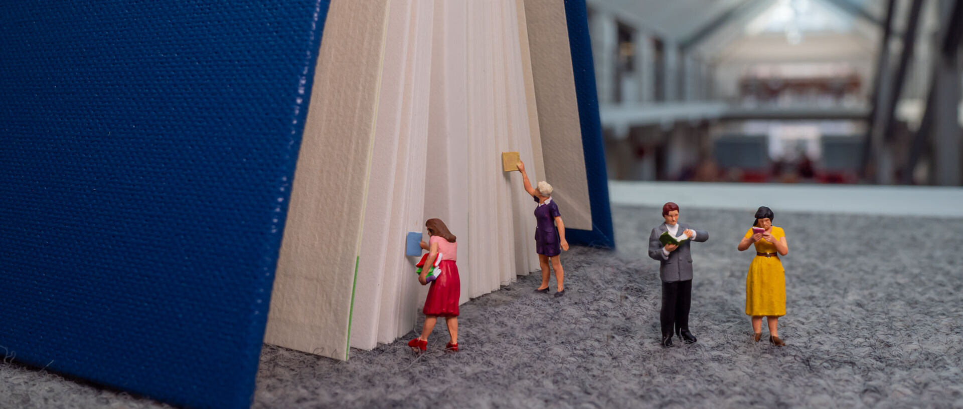 Zu erkennen ist ein großes Buch, zwei Miniaturfiguren lesen im Vordergrund, zwei weitere geben Bücher zurück oder lehen diese aus.