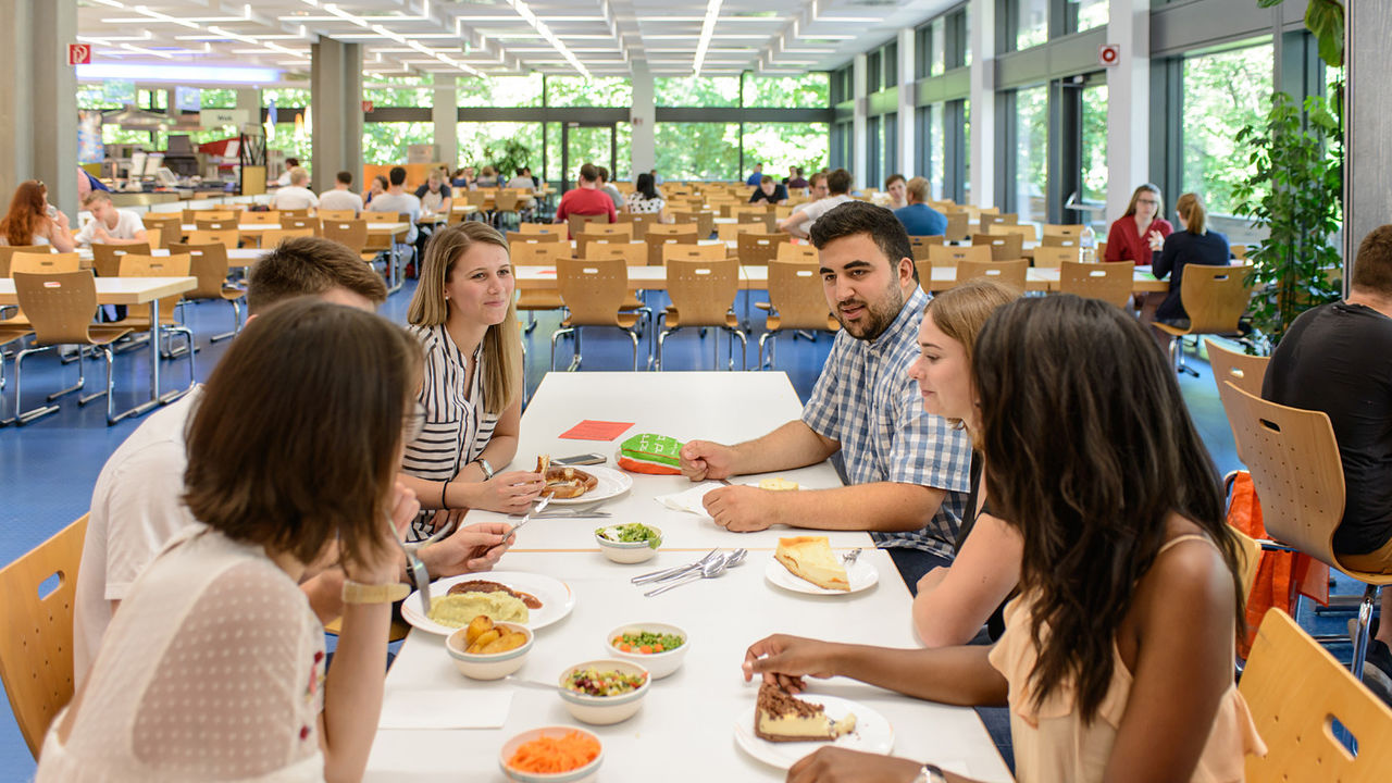 Studierende sitzen gemeinsam in der Mensa an einem Tisch und essen zu Mittag.