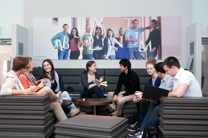 Acht Studierende sitzen in Lounge-Bereich im Gespräch, im Hintergrund Wandbild der Initiative Absolventum.