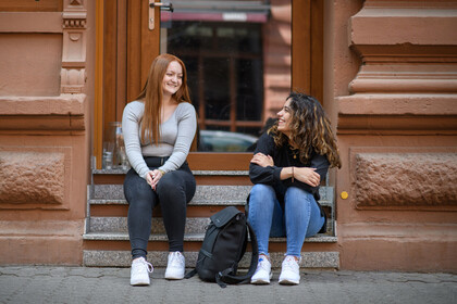 Zwei weiblich gelesene Studierende sitzen auf einer Treppenstufe in der Stadt.