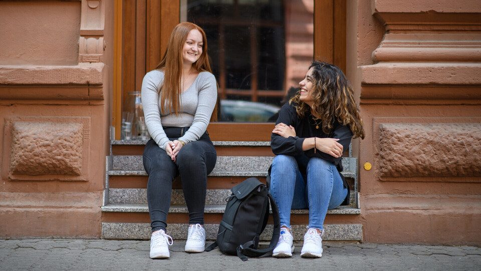 Zwei weiblich gelesene Studierende sitzen auf einer Treppenstufe in der Stadt.