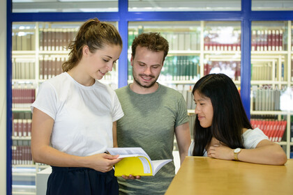 Drei Studierende befinden sich in der Unibibliothek und schauen gemeinsam in ein Buch.