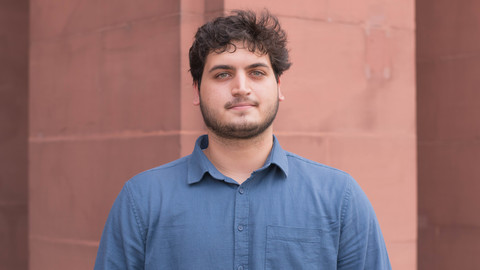 Ayman Tibi. He is wearing a blue shirt and is standing in front of a brown wall.