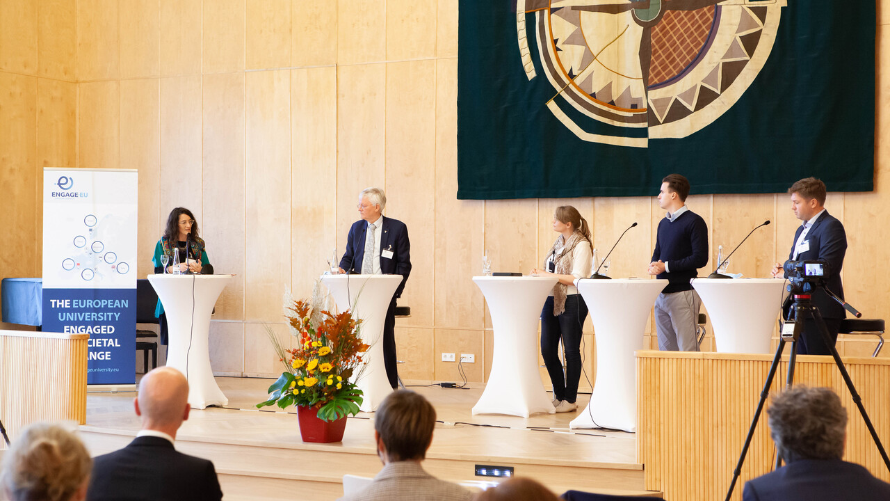 In einem Vorlesungssaal stehen fünf Personen in schicker Kleidung an jeweils einem Stehtisch. Sie stehen vor einem Publikum, das auf Stühlen sitzt.