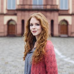 Juliane Koch trägt ihr rotes langes welliges Haar offen. Sie trägt eine rote Strickjacke und ein graues Oberteil. 