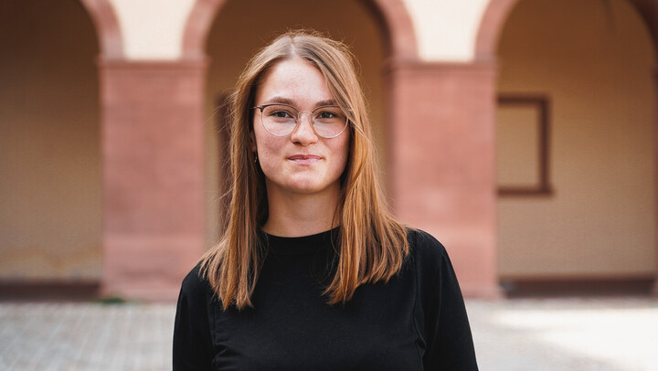 Johanna Mehltretter. She is wearing a black sweater and is standing in front of the Arcades of the castle.