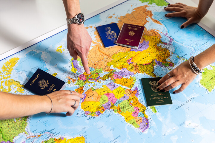 A map of the world lies on a table. Hands point to different places on it. Various passports are spread across the map.