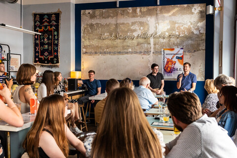 Vortrag mit Publikum in einer Bar, ein Mann spricht ins Mikrofon.