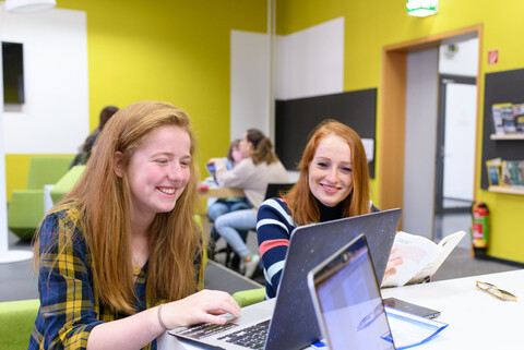 Zwei weiblich gelesene Studierende mit roten, langen Haaren sitzen an einem Tisch im Gruppenbereich der Schneckenhofbibliothek. Sie schauen lächelnd in einen Laptop vor ihnen, im hintergrund sitzen weitere Studierende..