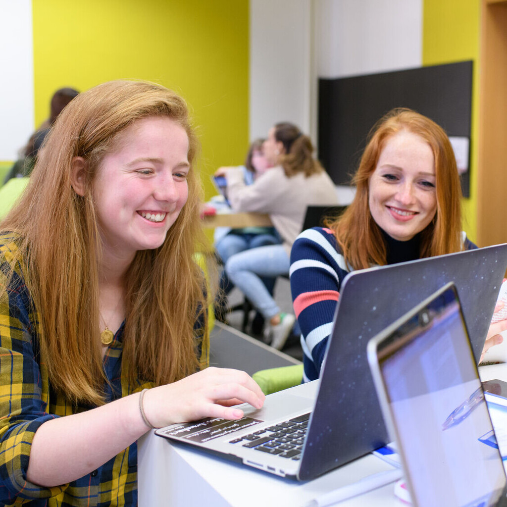Zwei weiblich gelesene Studierende mit roten, langen Haaren sitzen an einem Tisch im Gruppenbereich der Schneckenhofbibliothek. Sie schauen lächelnd in einen Laptop vor ihnen, im hintergrund sitzen weitere Studierende..