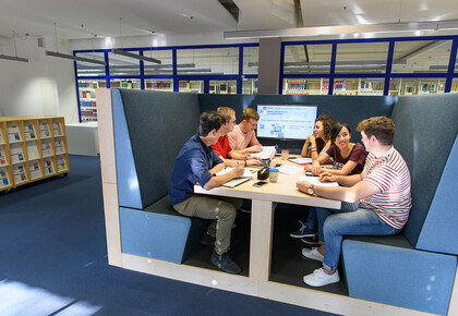 Eine Gruppe von Studierenden sitzt an einem U-förmigem blauen Sofa um einen Tisch. Am Ende des Tisches ist ein Bildschirm.