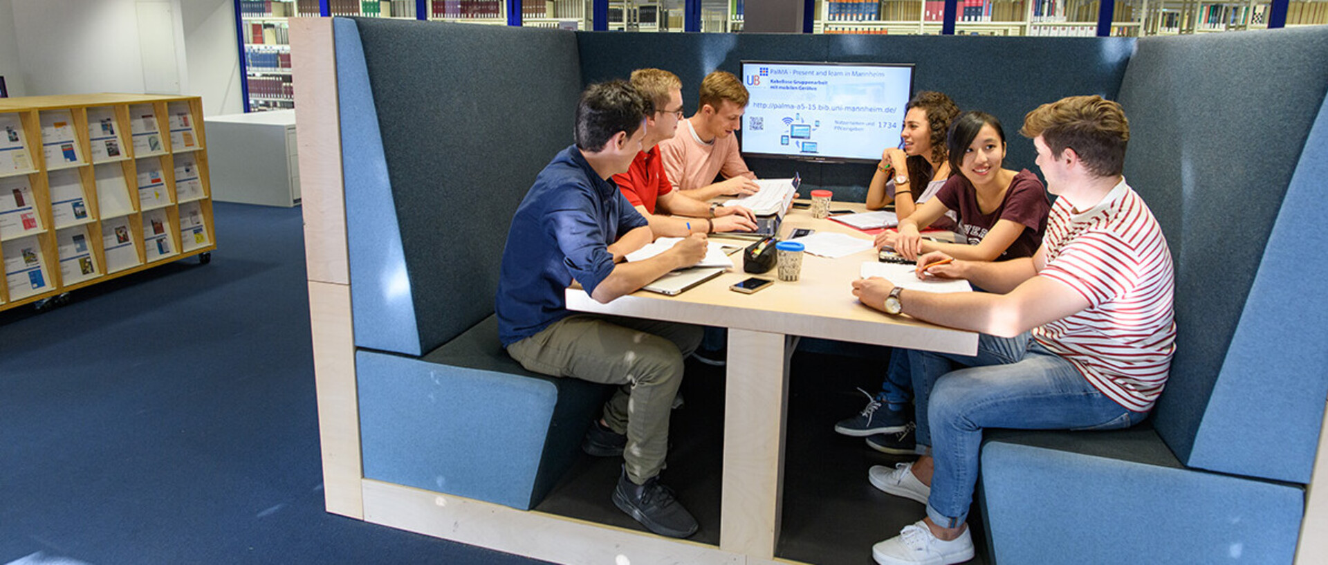 Eine Gruppe von Studierenden sitzt an einem U-förmigem blauen Sofa um einen Tisch. Am Ende des Tisches ist ein Bildschirm.