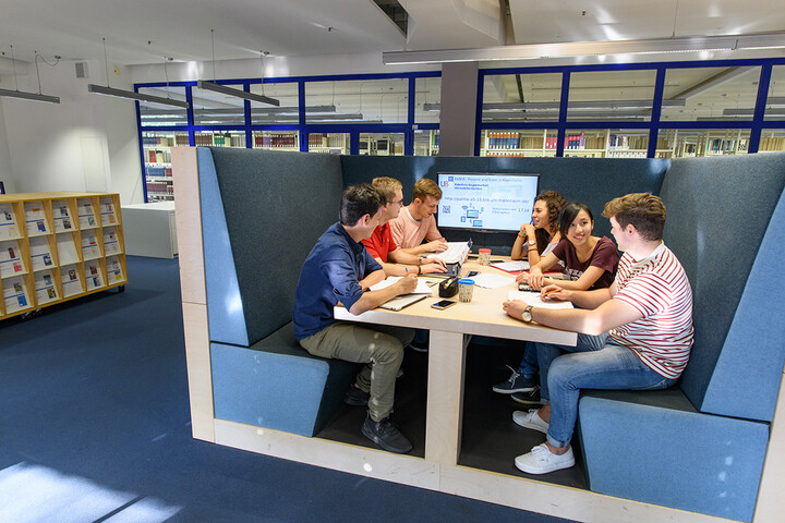 Eine Gruppe von Studierenden sitzt an einem U-förmigem blauen Sofa um einen Tisch. Am Ende des Tisches ist ein Bildschirm. 