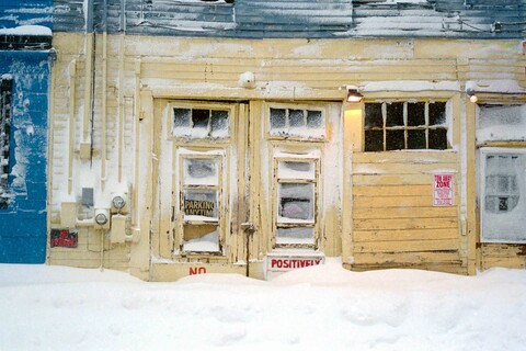 Eine Reihe schneebedeckter Häuserfronten. Am mittleren Haus hängt ein Coca-Cola-Ladenschild, am linken Haus ein Poster eines Mannes. 