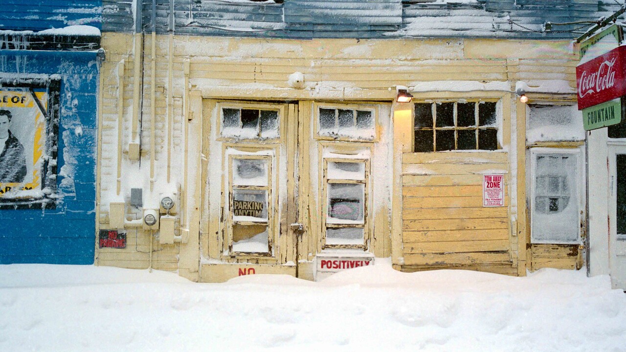 Eine Reihe schneebedeckter Häuserfronten. Am mittleren Haus hängt ein Coca-Cola-Ladenschild, am linken Haus ein Poster eines Mannes.