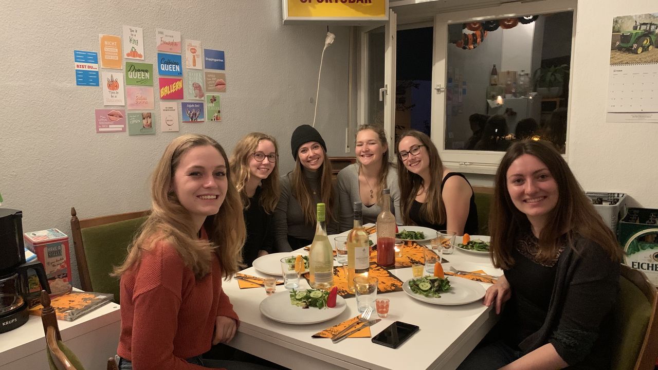 Sechs Studentinnen essen gemeinsam zu Abend. Über ihnen hängt eine Girlande aus schwarz-orangenen Kürbisgesichtern.