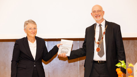 Zwei lächelnde Personen in schicker Kleidung stehen nebeneinander und halten die Festschrift zum 75-jährigen Jubiläum der Neubegründung in den Händen. Die Personen heißen Barbara Windscheid und Thomas Puhl.