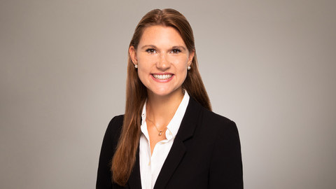 Victoria Meil. She is wearing a black suit and is standing in front of a brownish photowall.