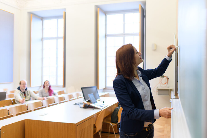 Eine Frau in blauem Jacket skizziert etwas auf einer Tafel. Im Hintergrund des Raums sitzen Studierende an Tischen.