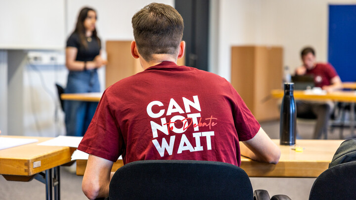 Eine Person mit kurzen Haaren trägt ein dunkelrotes Shirt mit der Aufschrift "Can not wait". Sie ist von hinten im Sitzen fotografiert.