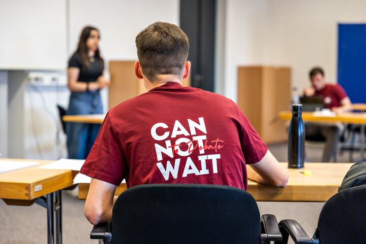 Eine Person mit kurzen Haaren trägt ein dunkelrotes Shirt mit der Aufschrift "Can not wait". Sie ist von hinten im Sitzen fotografiert.