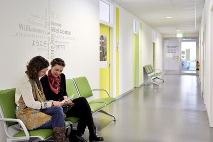 Two female students sit on a green upholstered bench in the corridor outside the Admissions Office and look at a leaflet. “Welcome” is written on the wall behind them in various languages.