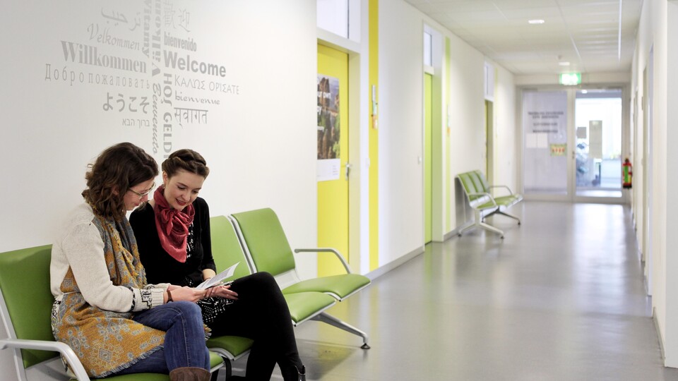 Zwei Studentinnen sitzen auf einer grünen gepolsterten Bank im Flur vor den Büros der Zulassungsstelle und sehen sich einen Flyer an. An der Wand hinter ihnen steht "Willkommen" in verschiedenen Sprachen.