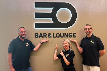 Drei Mitarbeitende stehen vor dem neuen Logo des Café EO. Sie tragen schwarze T-Shirts ebenfalls mit Logo, lächeln und zeigen auf das Logo.