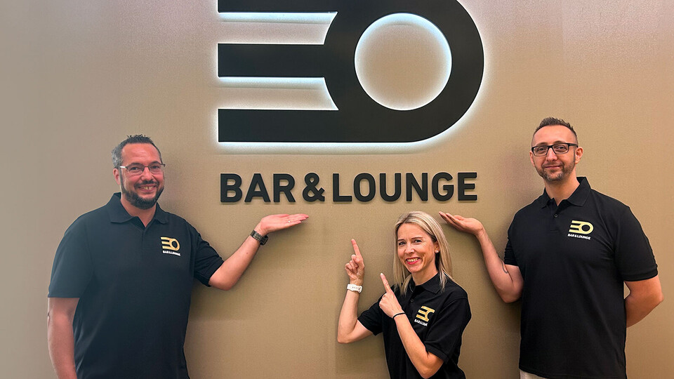 Drei Mitarbeitende stehen vor dem neuen Logo des Café EO. Sie tragen schwarze T-Shirts ebenfalls mit Logo, lächeln und zeigen auf das Logo.
