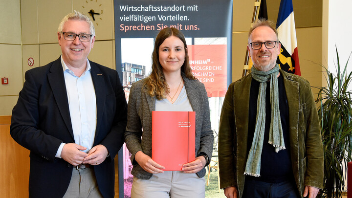 Drei Personen stehen nebeneinander, in der Mitte Laura Schmitt. Sie hält ein rotes Buch in der Hand.