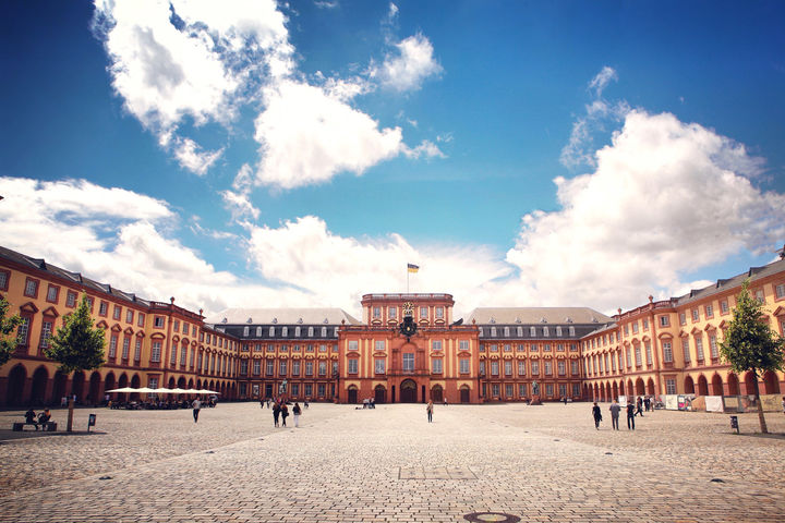 Das Barock-Schloss und der Ehrenhof der Universität Mannheim unter strahlend blauem Himmel. Das Schloss ist von unzähligen Fenstern, rotem Sandstein und einer gelben Fassade geprägt.