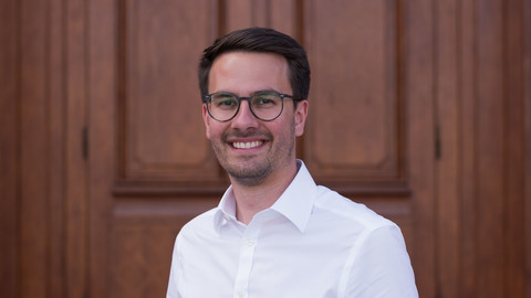 Steffen Zetzmann. He is wearing a white shirt and is standing in front of a brown door.