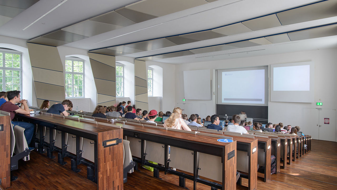 Studierende sitzen in einem Hörsaal und hören eine Vorlesung. Die Tische des Hörsaals sind aus dunklem Holz.