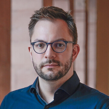 Sascha Adam hat kurze braune Haare und einen bart. Er trägt eine dunkle runde Brille und ein dunkelblaues Hemd.