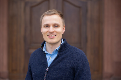 Er hat kurzes blondes Haar und trägt ein hellblaues Hemd unter einer dunkelblauen Weste.