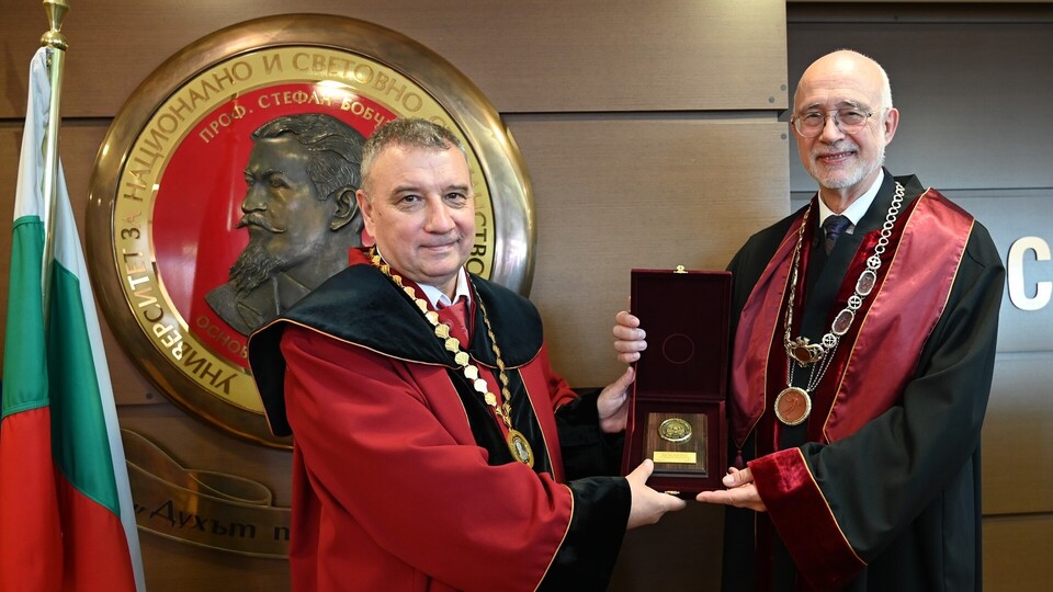 Zwei Männer in festlicher Robe - Professor Dimitar Dimitrov (links) und Professor Thomas Puhl - halten gemeinsam eine Medaille in einer Box, hinter ihnen ein großes Emblem und die bulgarische Flagge.