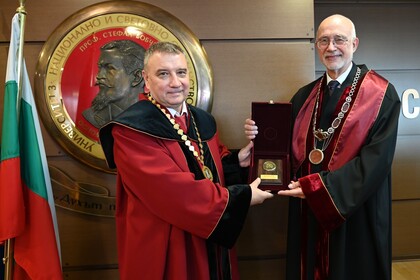 Zwei Männer in festlicher Robe - Professor Dimitar Dimitrov (links) und Professor Thomas Puhl - halten gemeinsam eine Medaille in einer Box, hinter ihnen ein großes Emblem und die bulgarische Flagge.