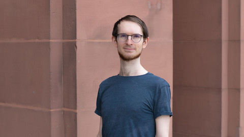 Frederic Gerdon. He is wearing a grey T-shirt and is standing in front of a brown stonewall.