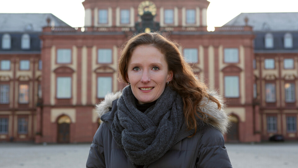 Portrait von Karin Hoisl. Sie trägt eine schwarze Jacke und einen grauen Schal, im Hintergrund ist das Mannheimer Schloß.