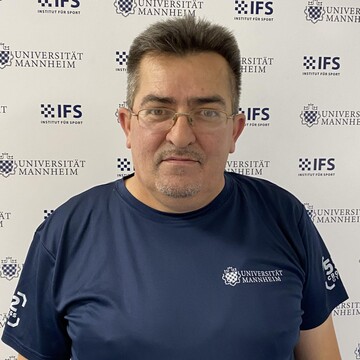 Sinan Seferaj hat kurze dunkle Haare. Er trägt eine schmale Brille und ein dunkelblaues Oberteil mit der Aufschrift "Universität Mannheim".