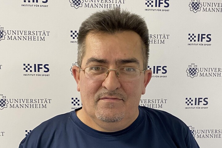 Sinan Seferaj hat kurze dunkle Haare. Er trägt eine schmale Brille und ein dunkelblaues Oberteil mit der Aufschrift "Universität Mannheim".