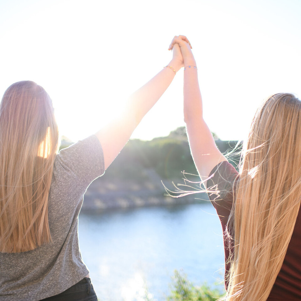 Zwei junge Frauen sind von hinten zu sehen. Sie schauen auf einen Fluss und reichen sich in der Luft die Hand.